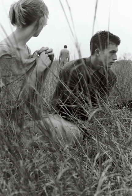 Monochrome portrait of a man and woman in a field of tall grass, with a third person walking in the distance.