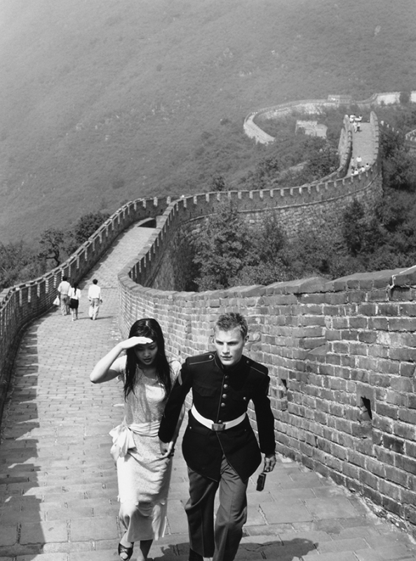 Monochrome shot of a couple walking hand-in-hand on the Great Wall of China, showcasing the wall's architecture and surrounding landscape.