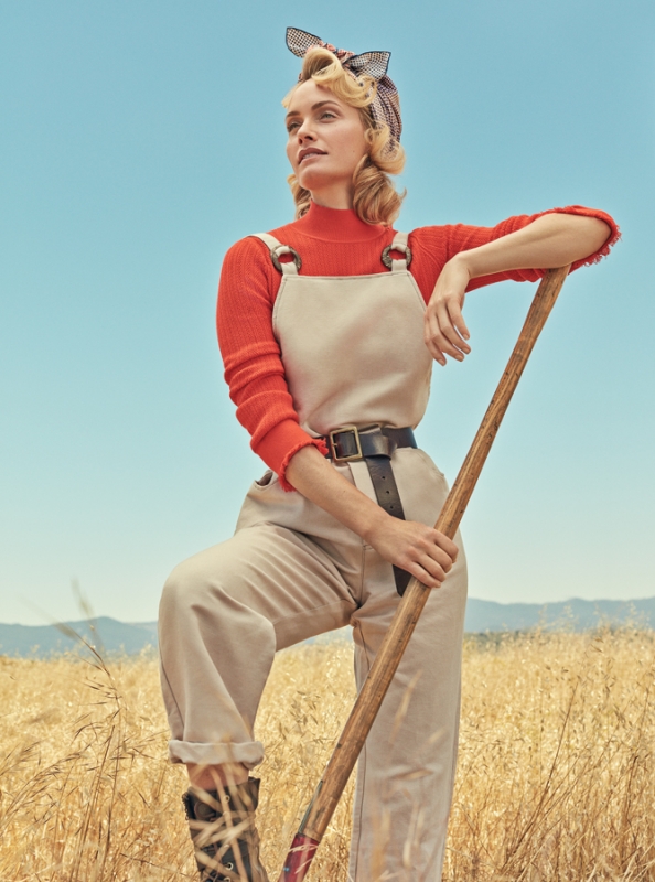 Blonde woman in overalls stands in wheat field with a shovel, smiling under clear blue sky. Retro vintage style photoshoot.