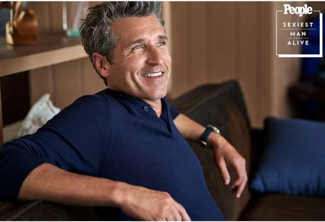 Patrick Dempsey smiling while seated on a dark sofa wearing a navy blue polo shirt.