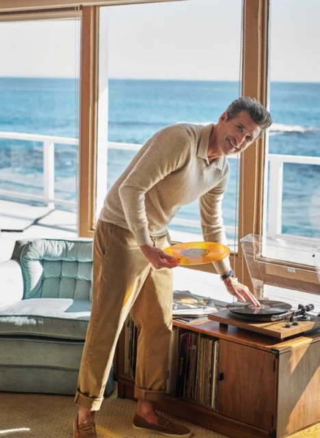 Adult male playing a vinyl record on a turntable inside a coastal home with ocean view, wearing casual clothing and smiling.