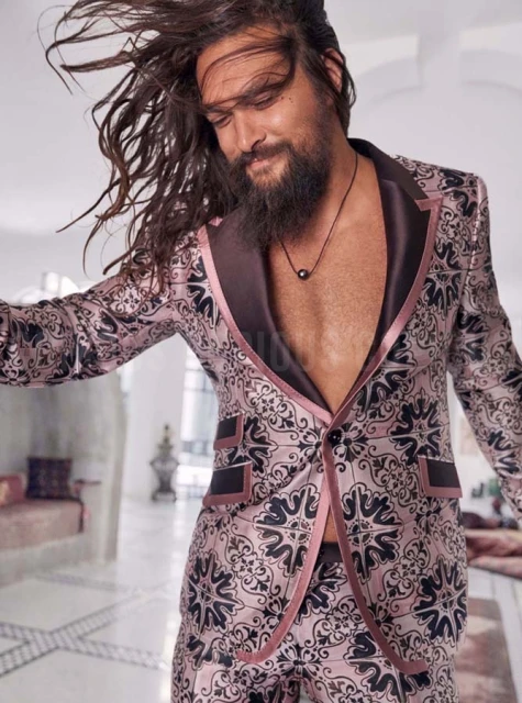 Portrait of Jason Momoa in a patterned pink suit with long hair, posing indoors with a light-colored floor and background.