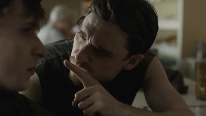 Close-up of two adult males indoors. One with a concerned expression points a finger, while the other listens intently in a dimly lit space.
