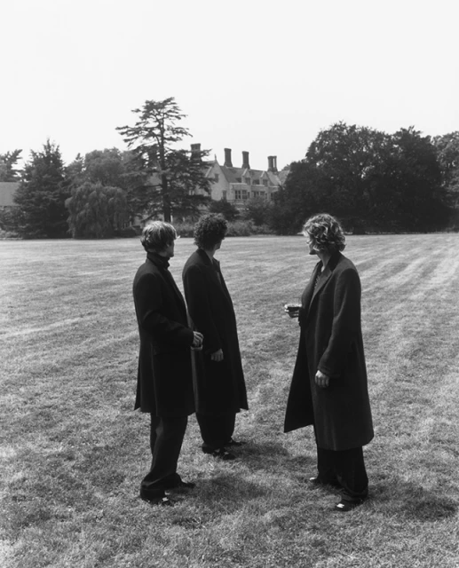 Three adult people in dark coats stand on a lawn in front of a large house, in a black and white photograph.