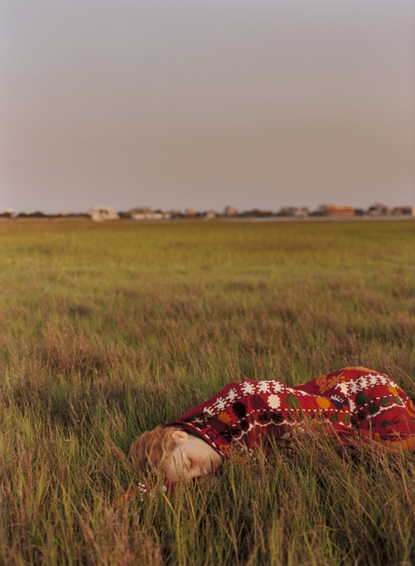 Woman lying down in tall green grass wearing a patterned red garment under an overcast sky