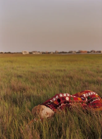 Woman lying down in tall green grass wearing a patterned red garment under an overcast sky