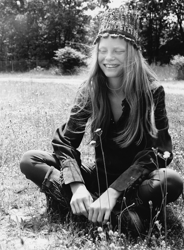 Monochrome portrait of a young woman in a natural setting, seated in a field, wearing a woven crown and a dark-coloured outfit.