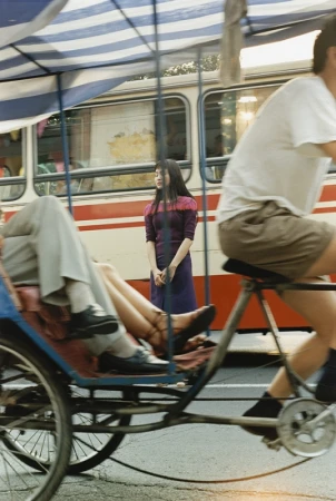 Woman in a purple dress stands near a bus in an urban scene, with a pedicab and cyclist in motion, capturing a moment of city life.