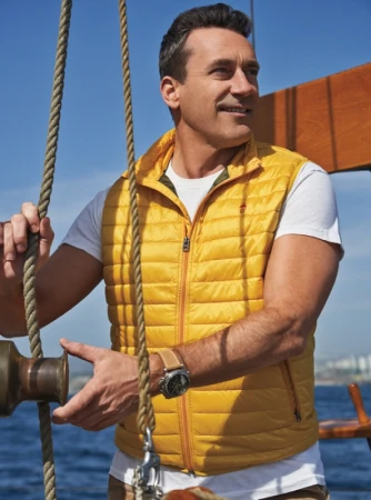 Man wearing a yellow quilted vest and white t-shirt on a sailboat with blue ocean in the background