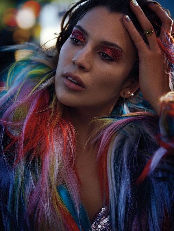 Close-up portrait of a woman with red glitter makeup and multi-colored fur coat, looking off-camera. The background is softly blurred.