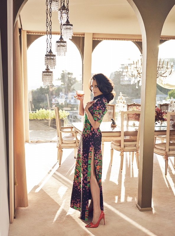 Elegant woman in patterned gown poses indoors holding a drink, lit by chandeliers, with a view of a cityscape outside large windows.