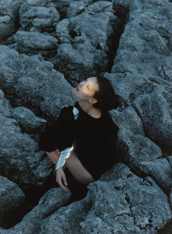 A woman in a black dress is sitting among grey rocks in a fashion editorial, gazing upwards with diffused sunlight on her face.