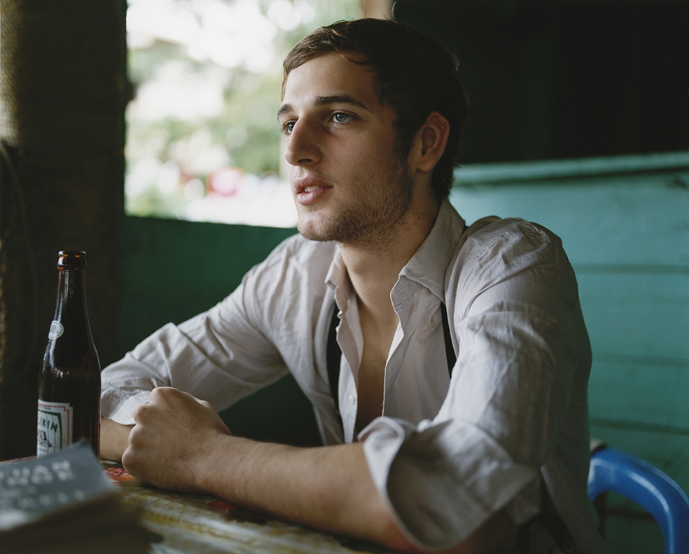 Young man with blue eyes sits at a table next to a beer bottle in an outdoor environment with a muted color palette and natural lighting.