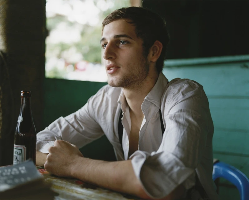Young man with blue eyes sits at a table next to a beer bottle in an outdoor environment with a muted color palette and natural lighting.