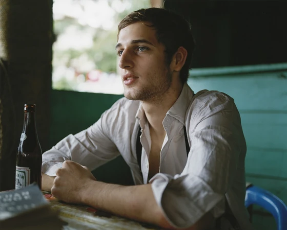 Young man with blue eyes sits at a table next to a beer bottle in an outdoor environment with a muted color palette and natural lighting.