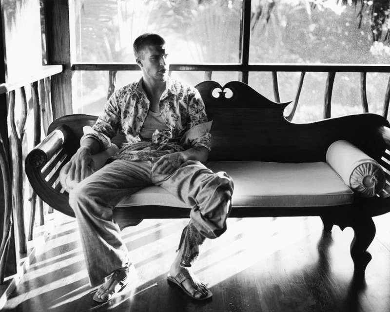 Black and white photo of an adult male model sitting on a wooden couch, looking away with natural light streaming through windows.