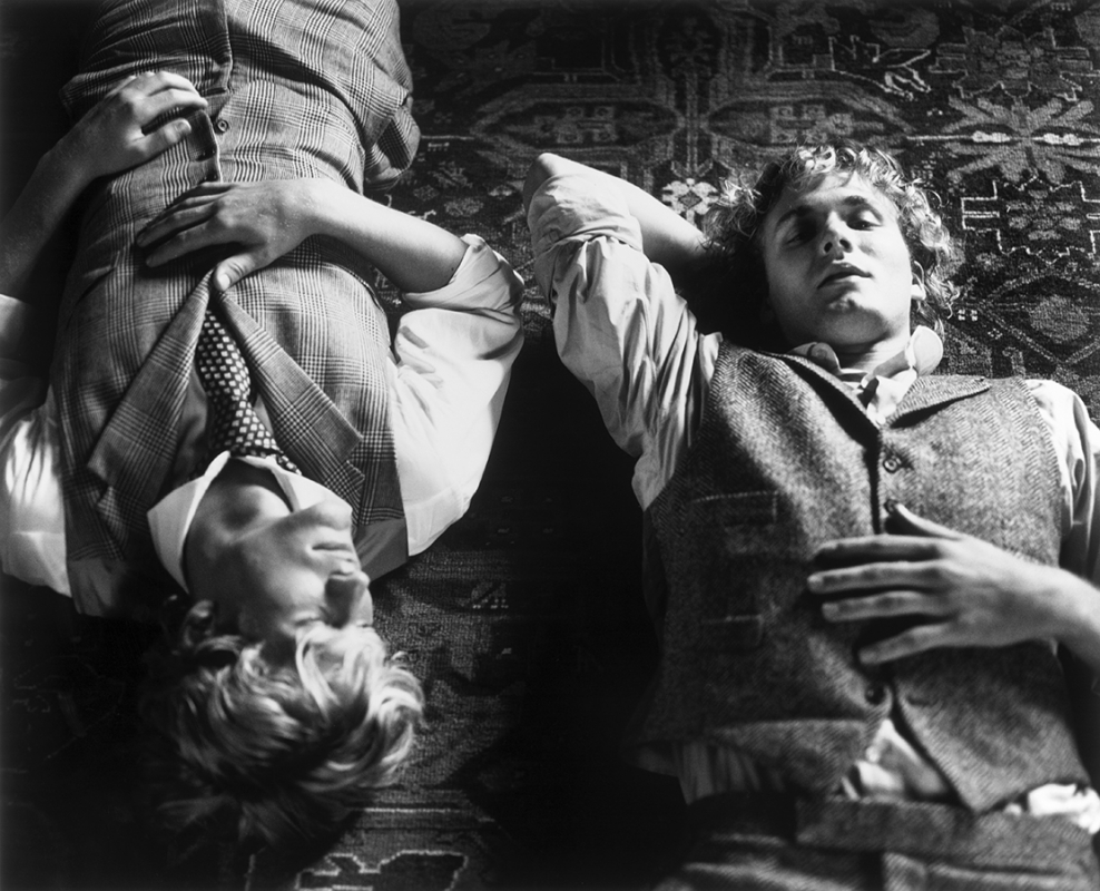 Monochrome portrait of two young adult males in vests lying on a patterned rug, looking relaxed in casual, classic menswear fashion.