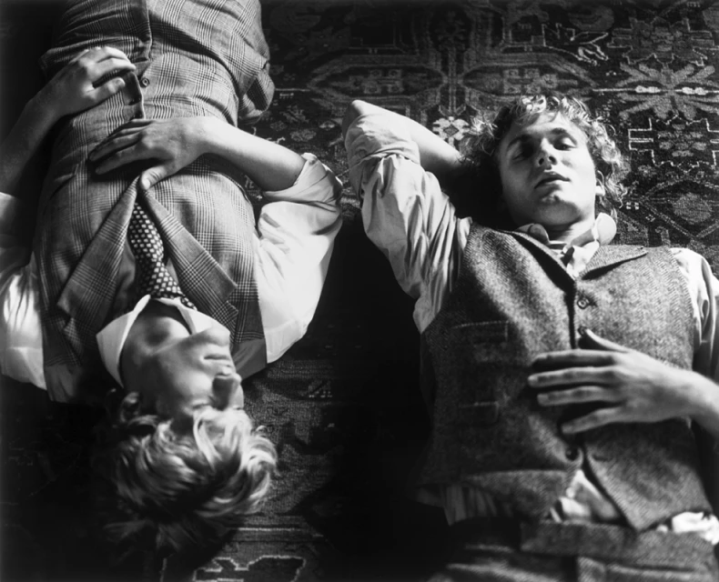 Monochrome portrait of two young adult males in vests lying on a patterned rug, looking relaxed in casual, classic menswear fashion.