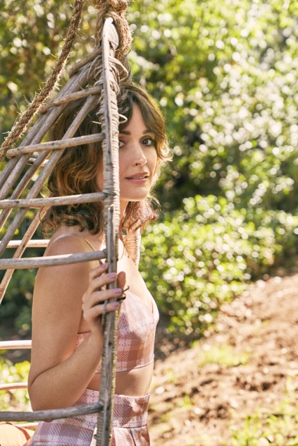 Portrait of a woman with wavy brown hair in a pink and white plaid outfit sitting in a wooden hanging chair in a green outdoor setting.