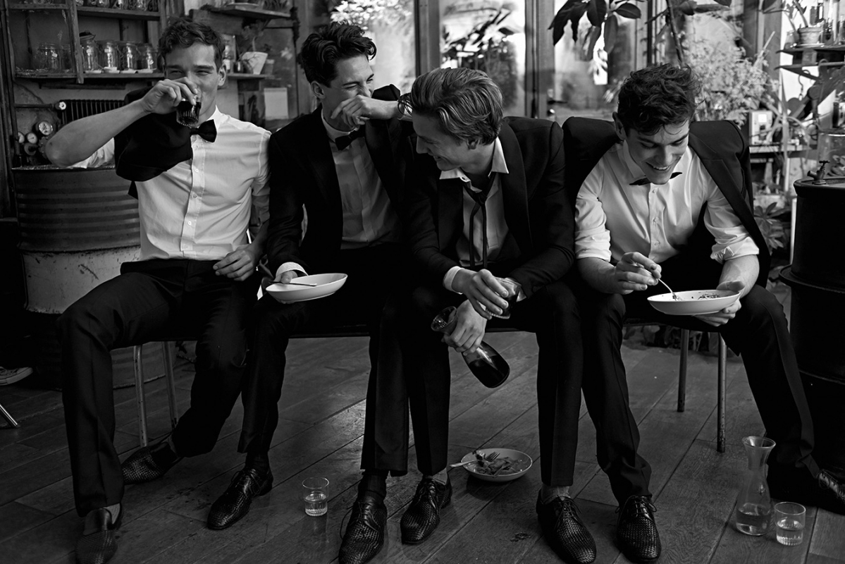 Four young men in formal wear sit closely together indoors, eating and drinking in a monochrome image that captures camaraderie and style.