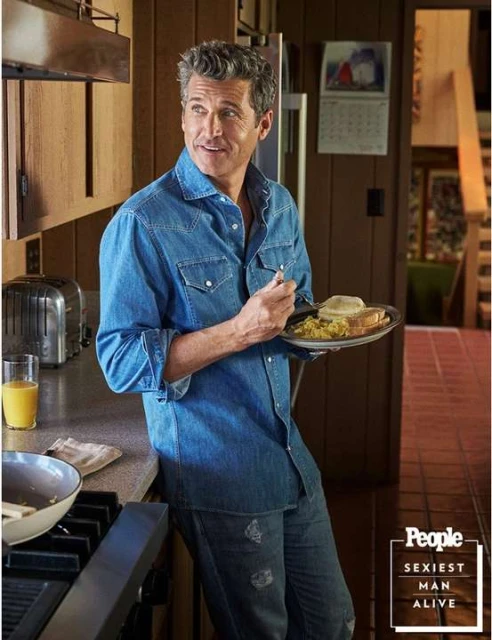 Adult man in denim stands in a kitchen holding a plate of food. The background features cabinets, a toaster, and natural light.