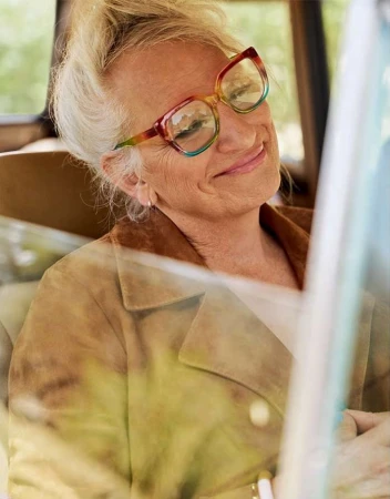 Elderly woman wearing colourful gradient glasses and tan jacket inside a car, photographed by Carter Smith