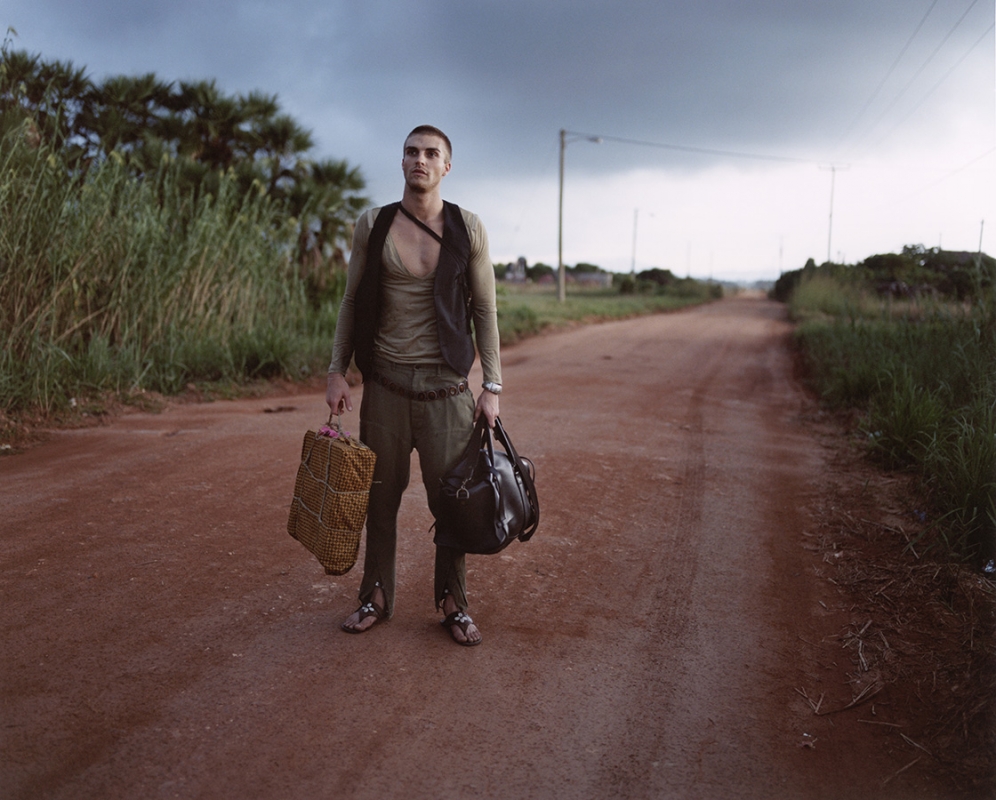 Male model poses with luggage on a dirt road, wearing an open shirt and vest, rural setting with storm clouds above.