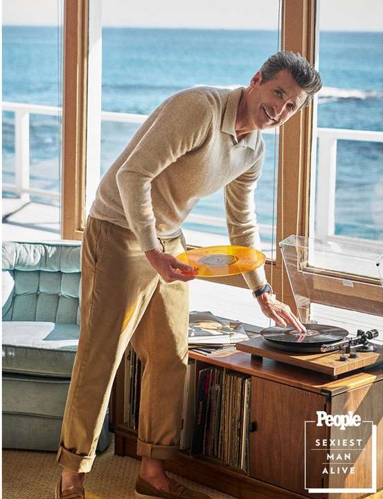 Patrick Dempsey shown indoors in front of ocean view window, wearing a sweater and khakis, placing an orange vinyl record on a turntable.