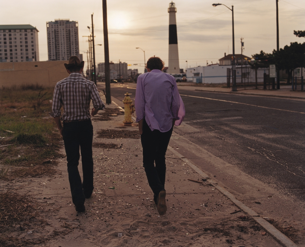 Two adult males are walking away from the viewer on a sidewalk in an urban setting with buildings and a lighthouse in the background.