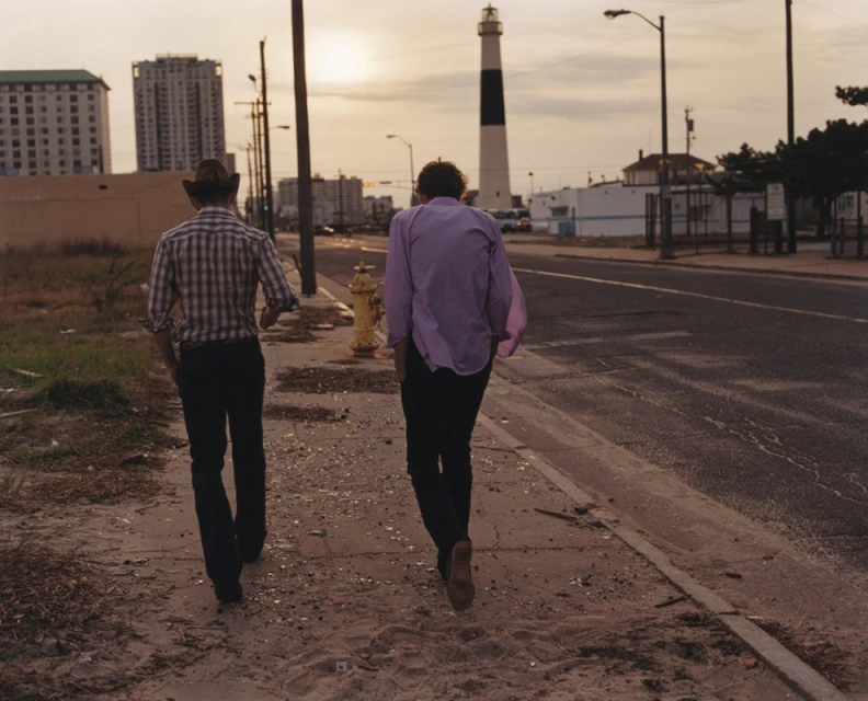 Two adult males are walking away from the viewer on a sidewalk in an urban setting with buildings and a lighthouse in the background.
