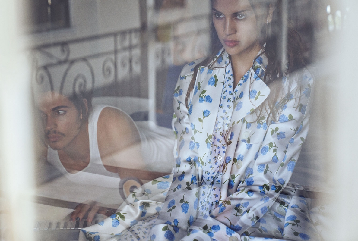 Young woman in floral shirt and man in white tank top indoors, viewed through a glass pane with reflections and soft, diffused lighting.