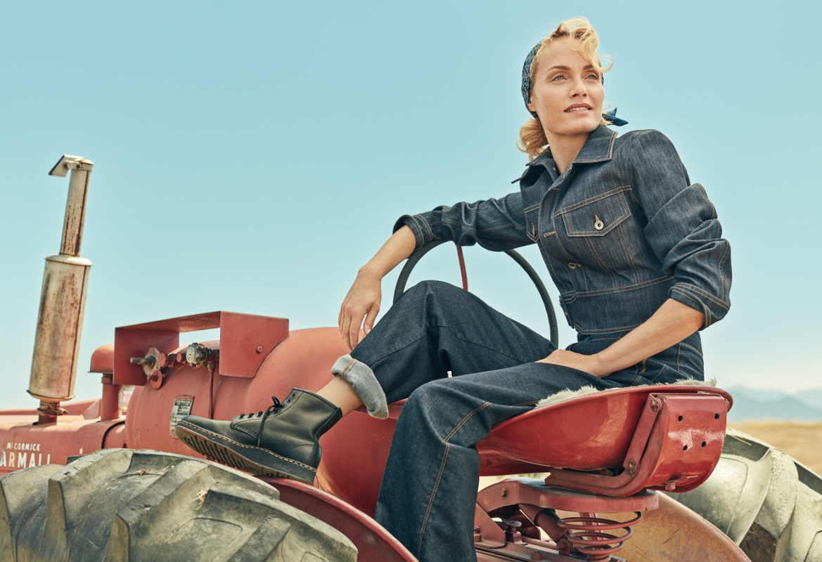 Adult female in a denim jumpsuit sits on a red tractor in a rural setting with blue sky. Black boots and a headscarf complete the look.
