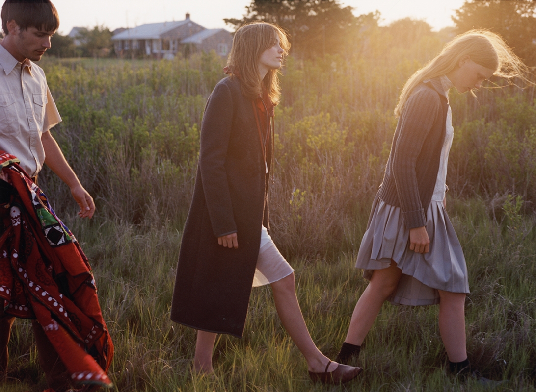 Three people walk through a grassy field at sunset, with one adult male and two young adult females in vintage-style clothing.