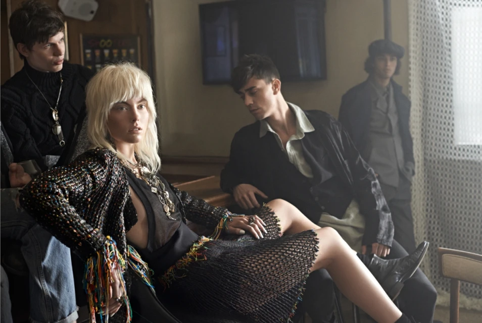 A group of four young adults in fashionable attire are indoors in a casual setting. One woman with blonde hair is seated with her legs visible.