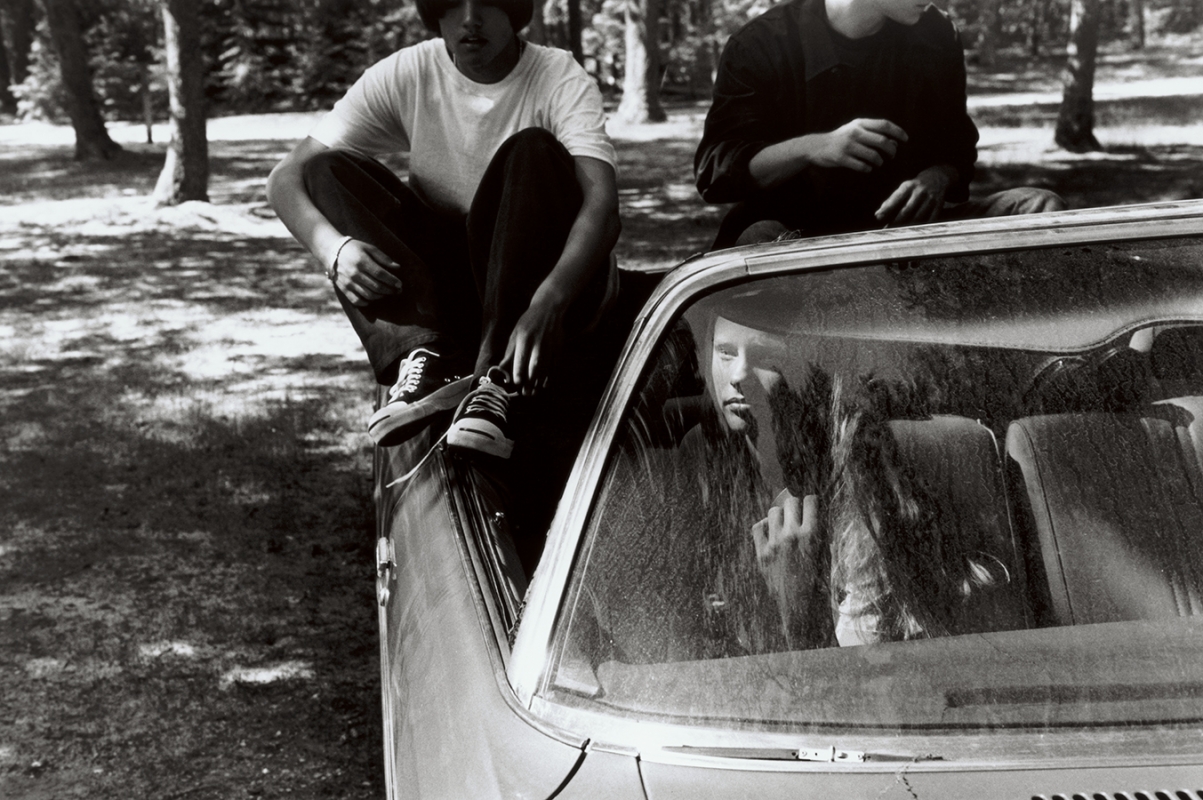Monochrome image of people interacting around a convertible car outdoors near trees with one person seated inside the vehicle.