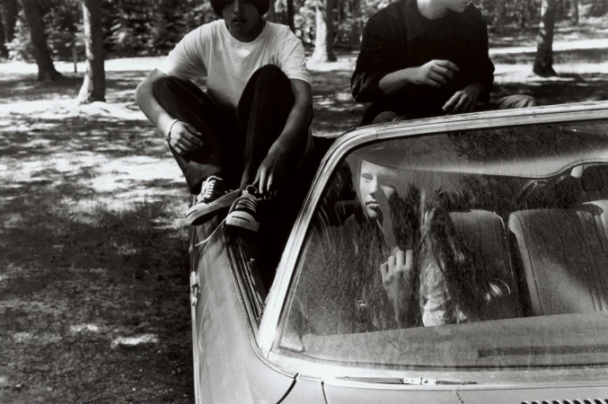 Monochrome image of people interacting around a convertible car outdoors near trees with one person seated inside the vehicle.