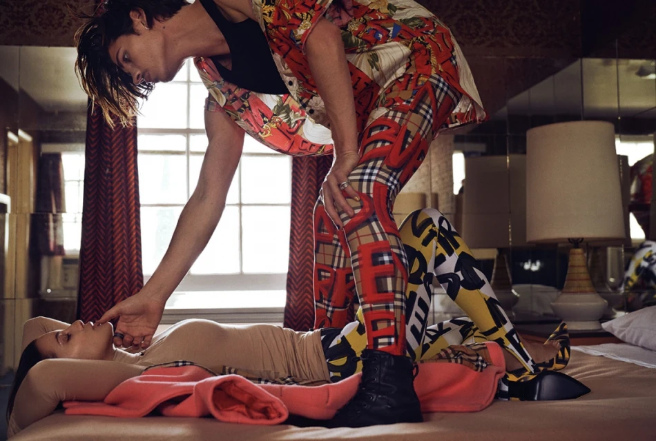 Two fashion models in a studio setting. One woman lies on a bed while another person stands over her. Both models wear colorful patterned outfits.
