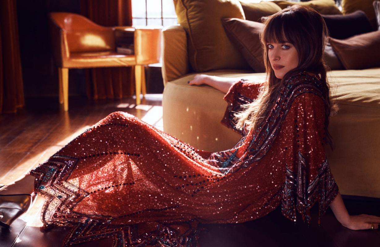 Woman in a sequined dress poses indoors, seated on the floor with furniture visible in the background, bathed in warm light.