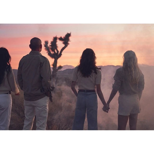 Family of four silhouetted against a desert sunset with pink and orange hues. The background features a Joshua tree and distant mountains.