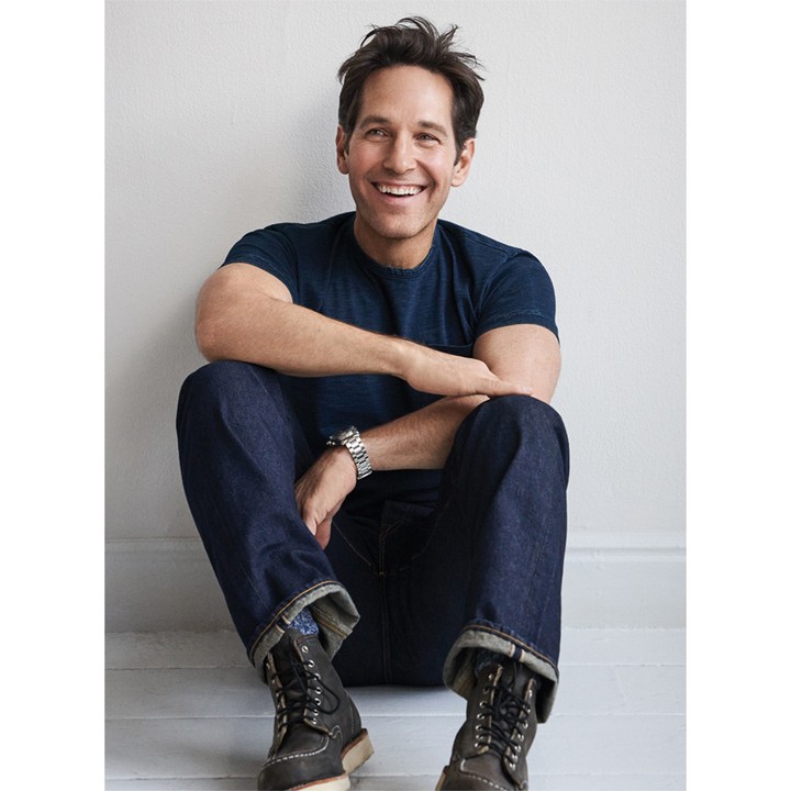 Portrait of Paul Rudd sitting against a white wall wearing dark blue t-shirt and jeans with boots