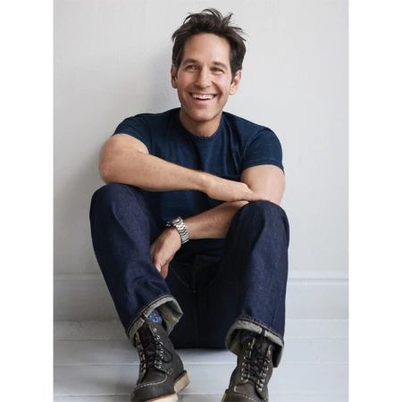 Portrait of Paul Rudd sitting against a white wall wearing dark blue t-shirt and jeans with boots