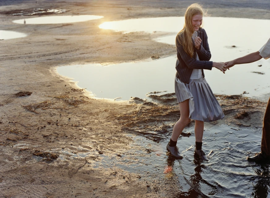 Young woman in gray skirt and cardigan smiles, holding hands with another person, walking through a shallow water landscape at sunset.