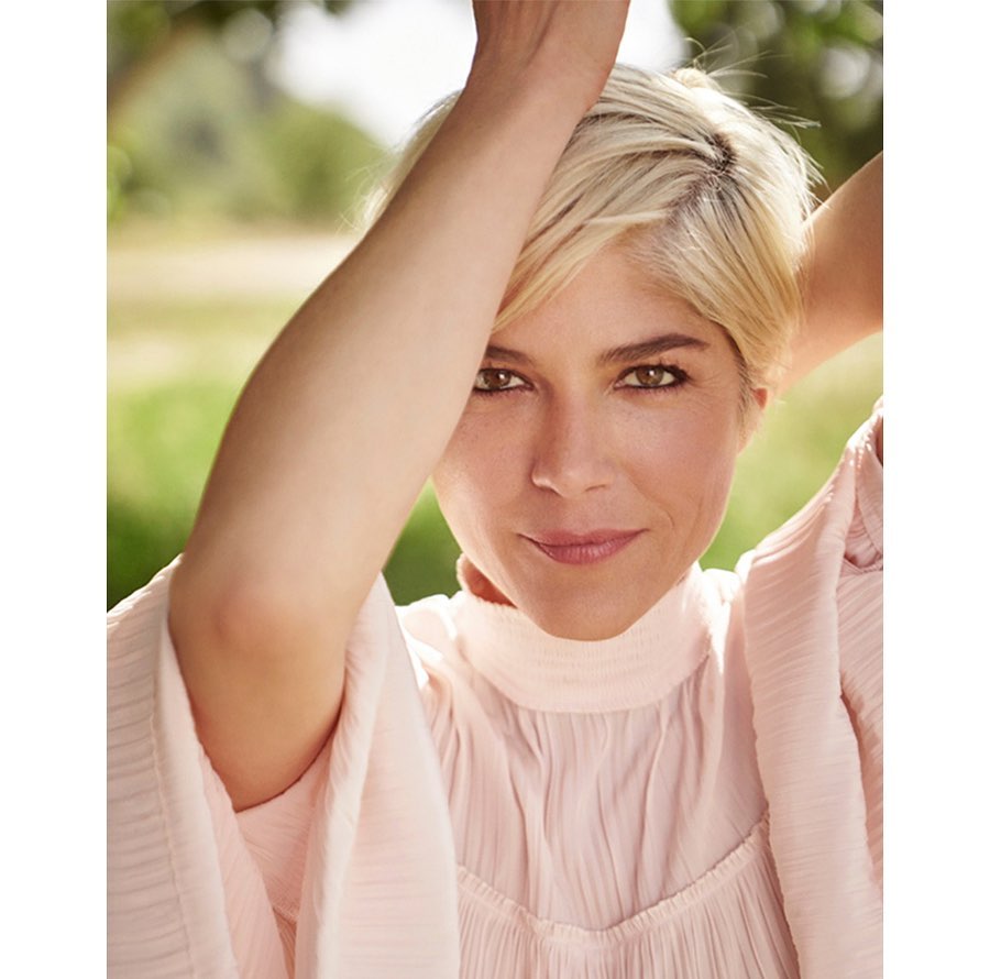 Close-up portrait of a smiling adult woman with blonde hair, wearing a light pink top, posed outdoors with green trees in the background.