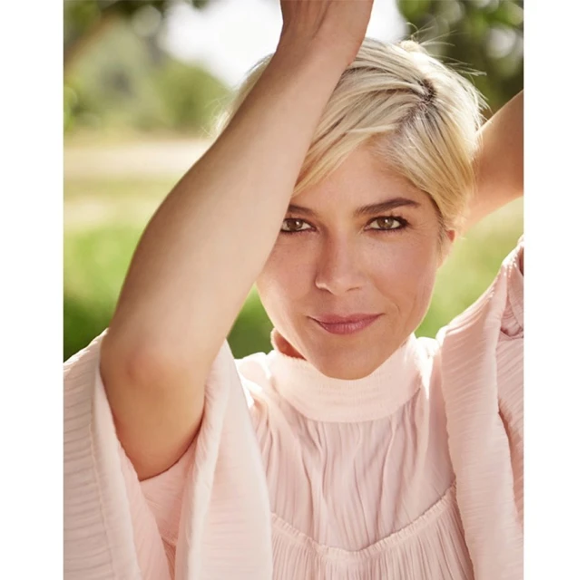 Close-up portrait of a smiling adult woman with blonde hair, wearing a light pink top, posed outdoors with green trees in the background.