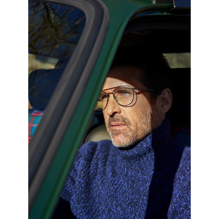 Adult male in green car wearing glasses and blue sweater, looking out the window on a sunny day. Automotive fashion editorial.