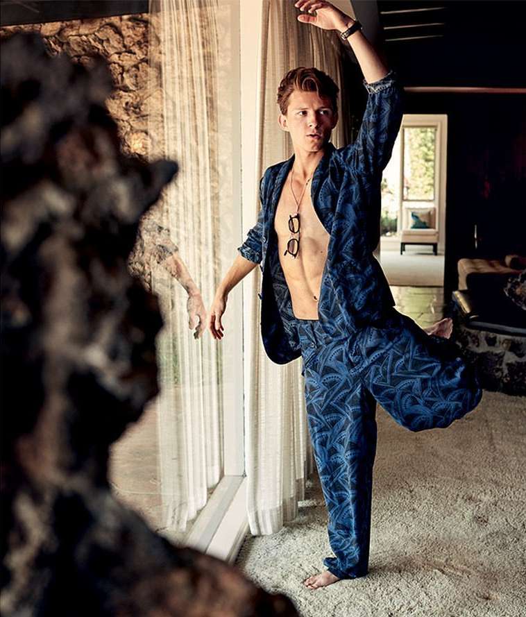 A Caucasian male in a patterned suit strikes a ballet pose indoors, with an open jacket displaying his torso, in a stylishly lit interior.