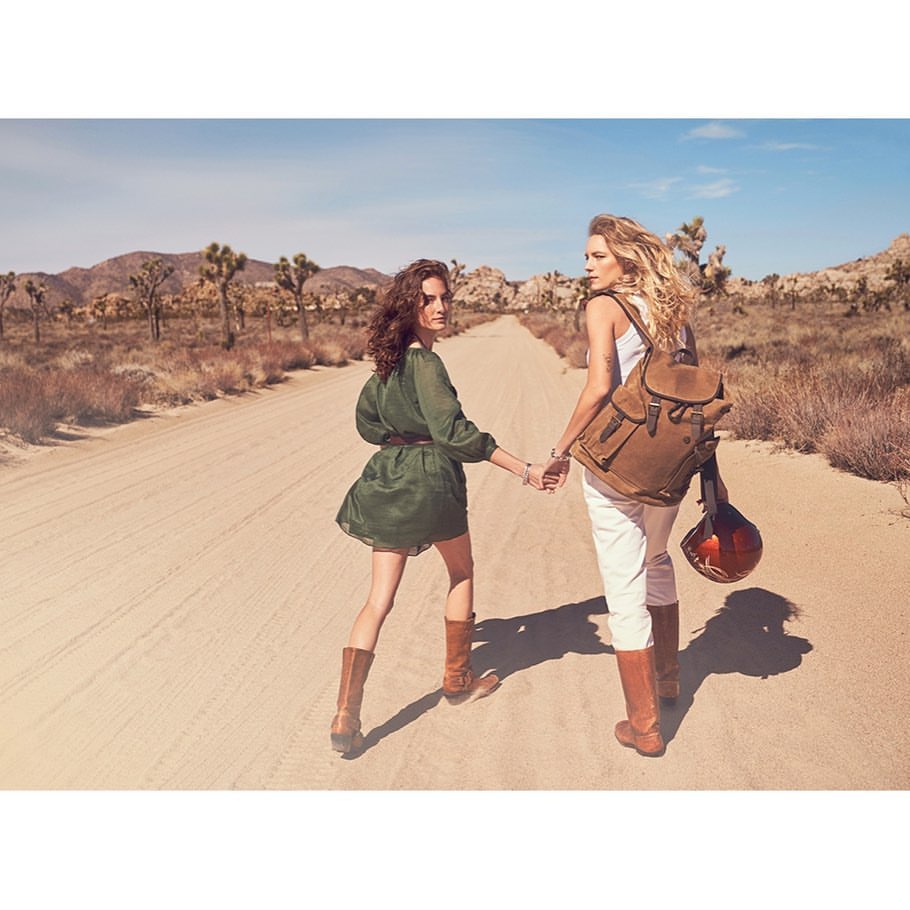 Two women walk on a desert road; one wears a backpack, the other a dress. They are holding hands with a desert landscape in the background.