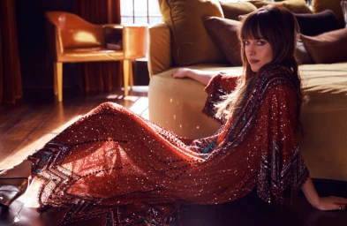 Dakota Johnson seated on a hardwood floor wearing a heavily embellished sequined red caftan dress.
