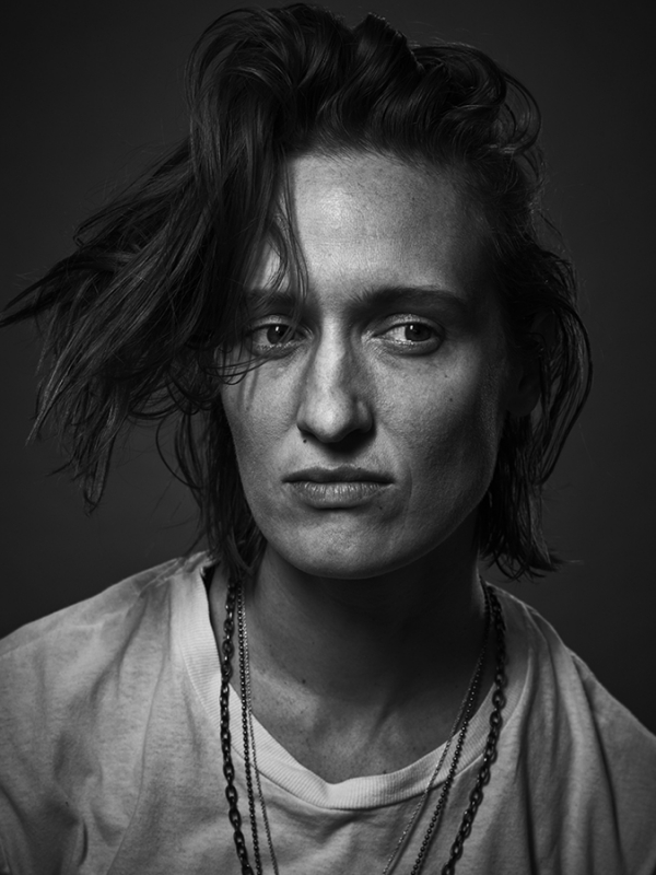 Monochrome portrait of a person with tousled hair wearing a white t-shirt and layered chain necklaces by Justin Stephens