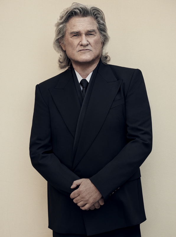 Portrait of Kurt Russell wearing a black tuxedo with silver hair, posing against a neutral-colored wall in a formal setting.
